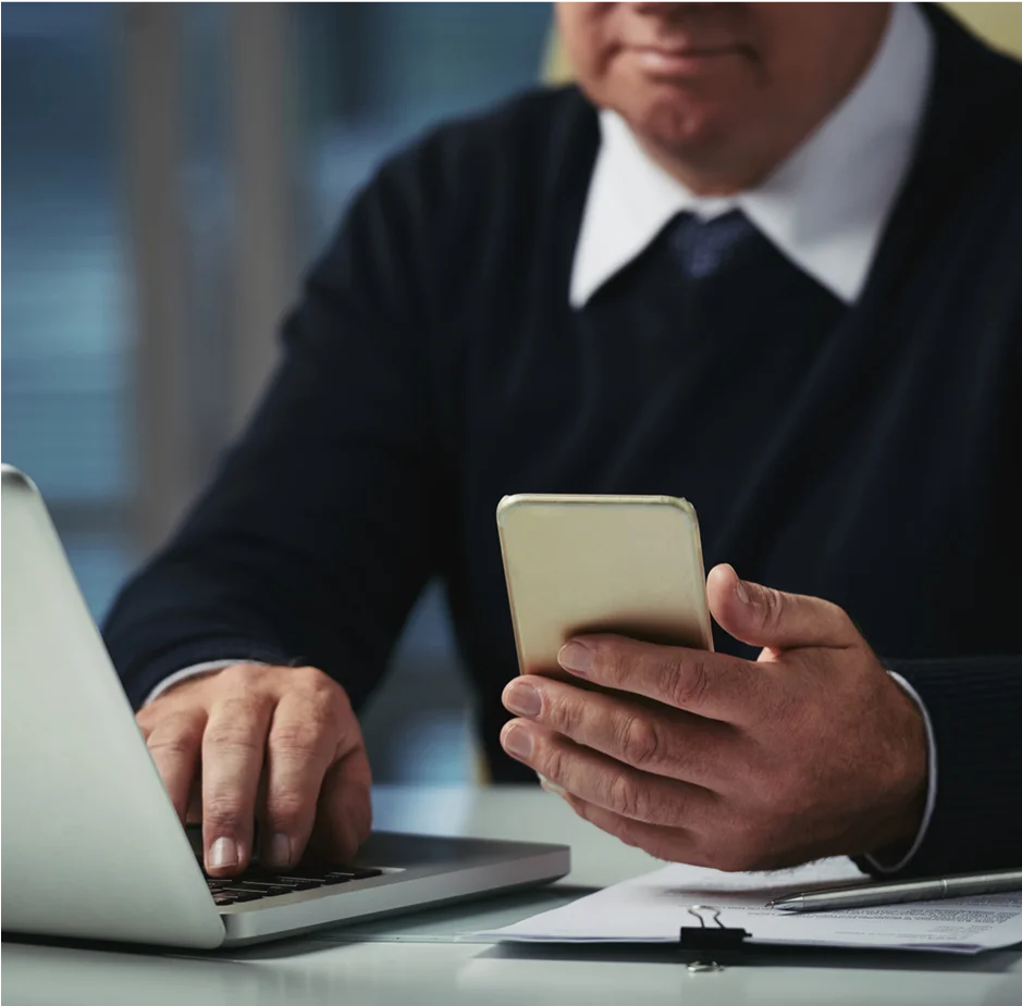 A man seated at a desk, working on a laptop with a cell phone beside him.
